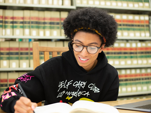 A student writes in a notebook at a desk in a library.